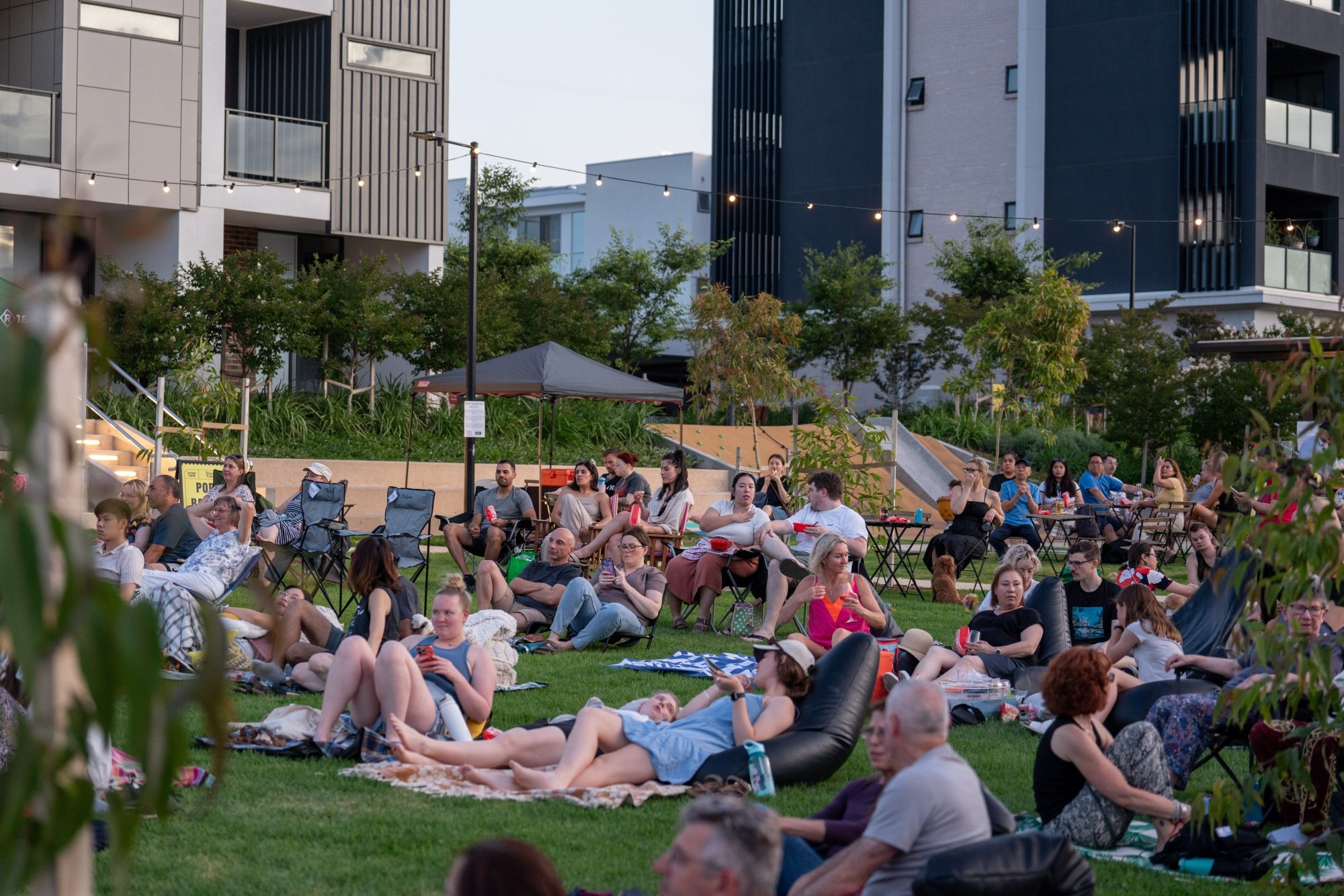 People sitting and lying on blankets and bean bags on a grassy area, enjoying an outdoor event surrounded by modern apartment buildings.