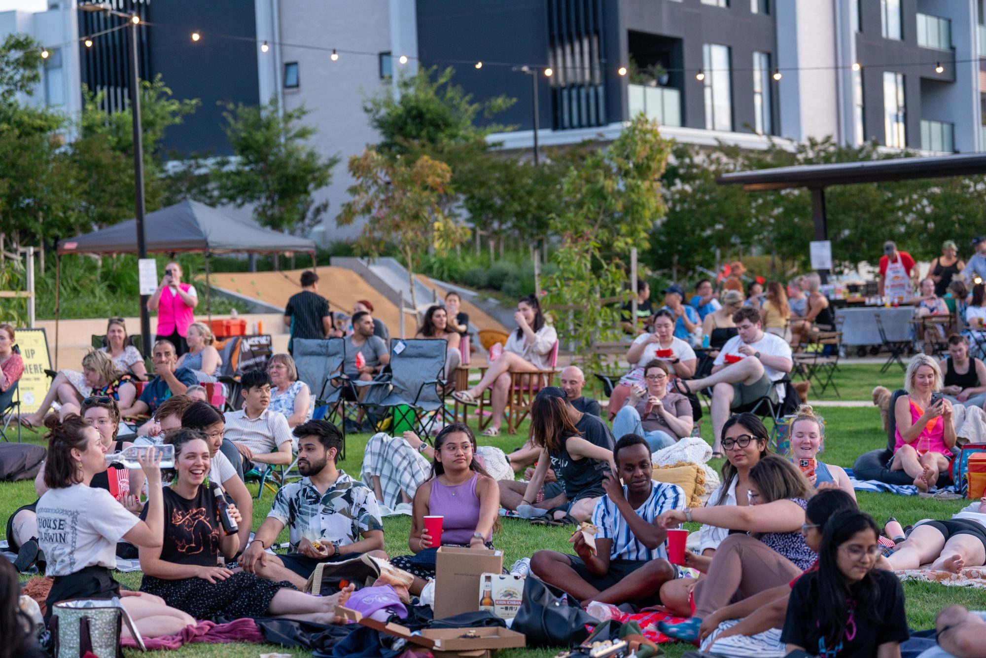 Close-up of a diverse group of people enjoying a relaxed outdoor community event on a lawn, seated on blankets and chairs with food and drinks, surrounded by string lights and modern apartment buildings.