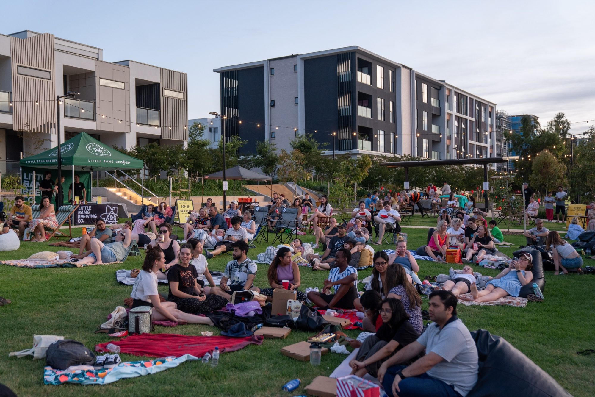Large crowd gathered on a grassy area, sitting on picnic blankets and bean bags at an outdoor community event, with modern apartment buildings and event tents in the background.