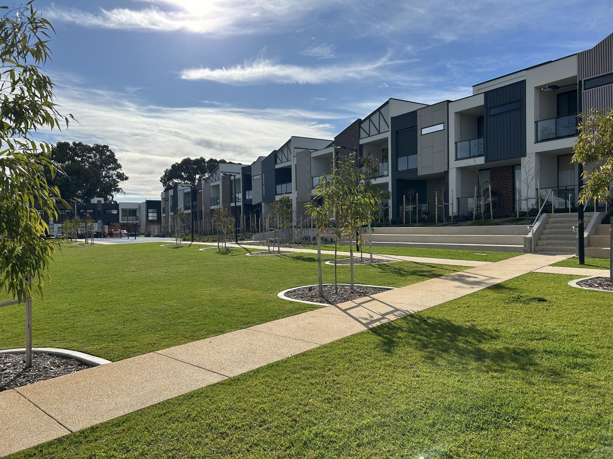 Modern green space in a residential area with a sunny blue sky, featuring young trees in mulched beds along a paved footpath and contemporary townhouses in the background.
