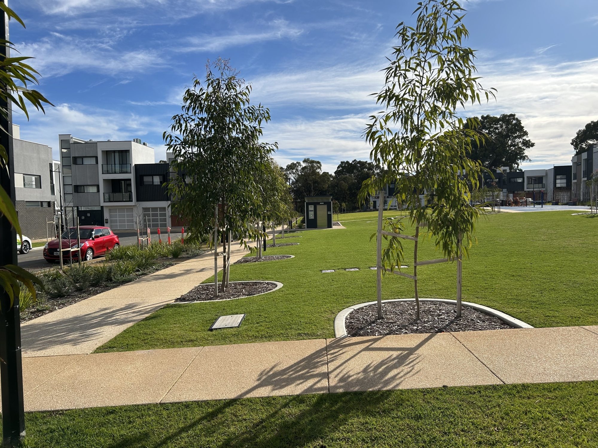 Sunny urban park with young trees planted along a curved paved pathway, surrounded by neatly trimmed grass and modern residential buildings.