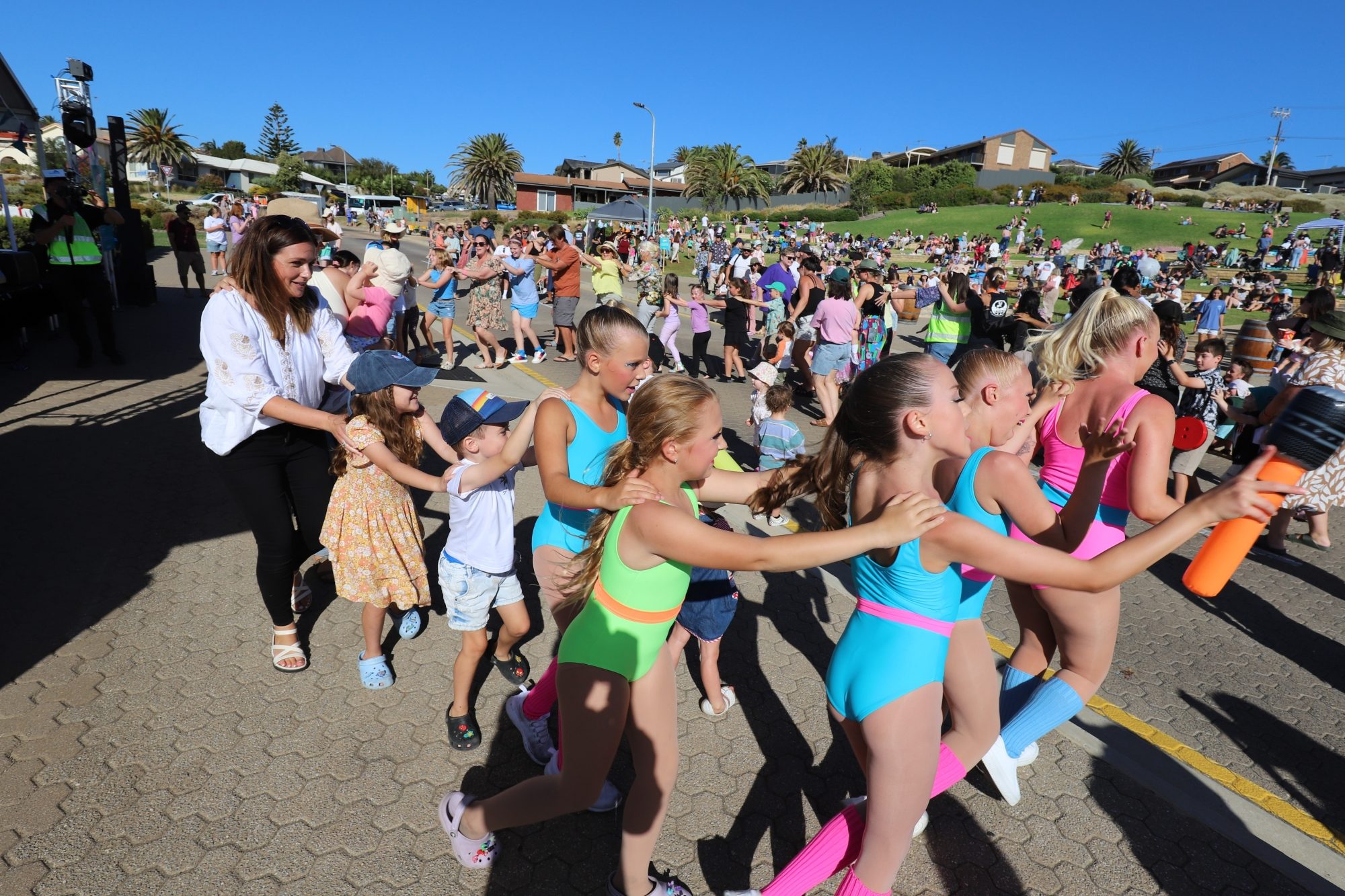 Outdoor community event with a large crowd gathered on a sunny day. A group of children in colourful dance costumes perform in a conga line, followed by parents and younger children. People are seated on grassy slopes in the background, enjoying the lively atmosphere.