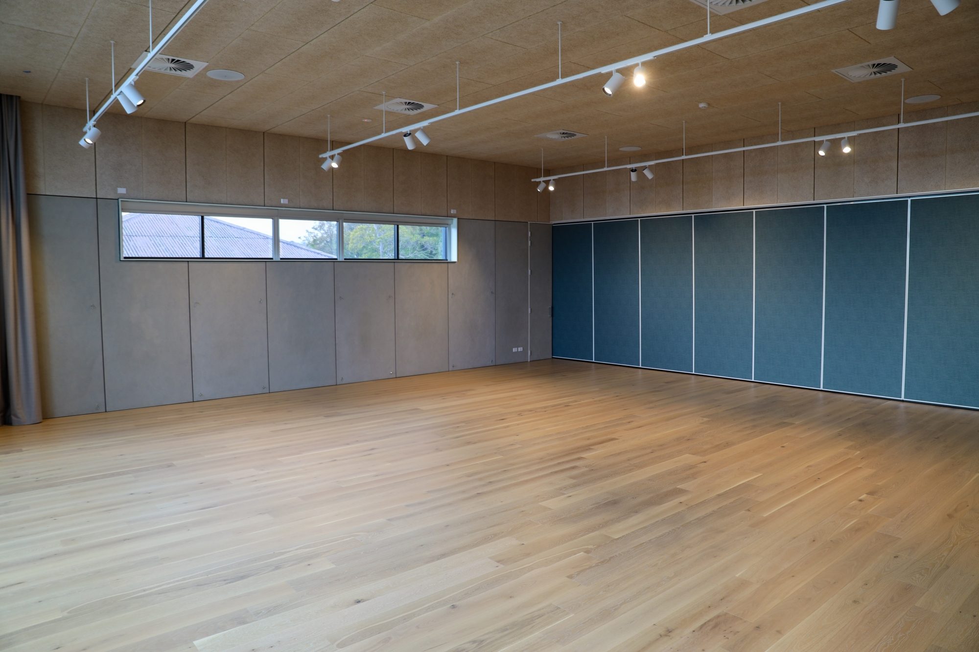 Large empty room with light wood flooring, grey walls, a small horizontal window, ceiling track lighting, and blue folding wall panels.
