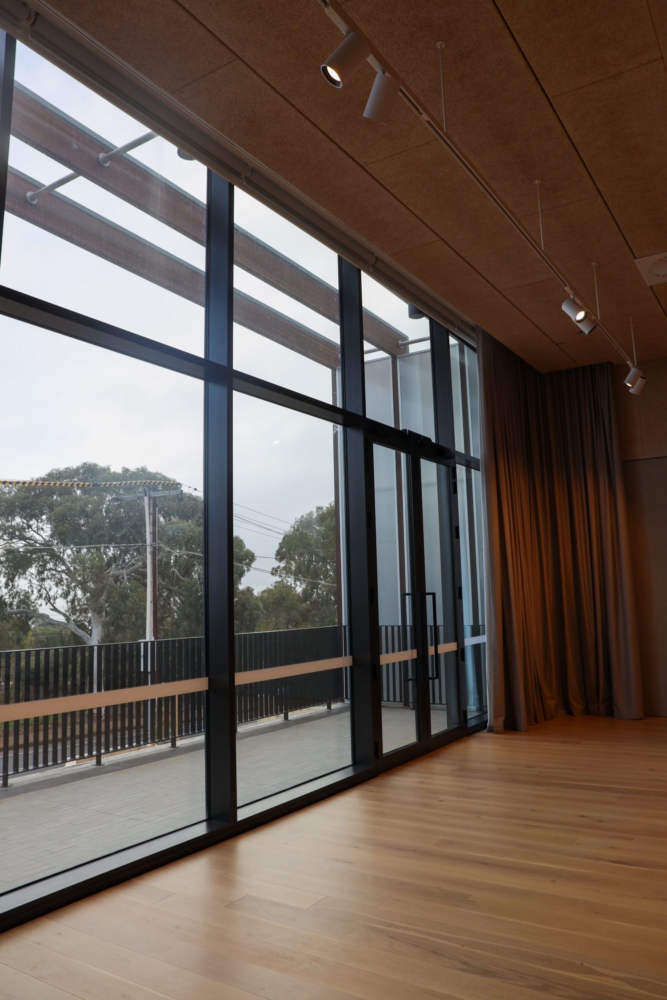 Spacious room with large floor-to-ceiling windows, wood flooring, and ceiling track lights, overlooking trees and an outdoor balcony area.