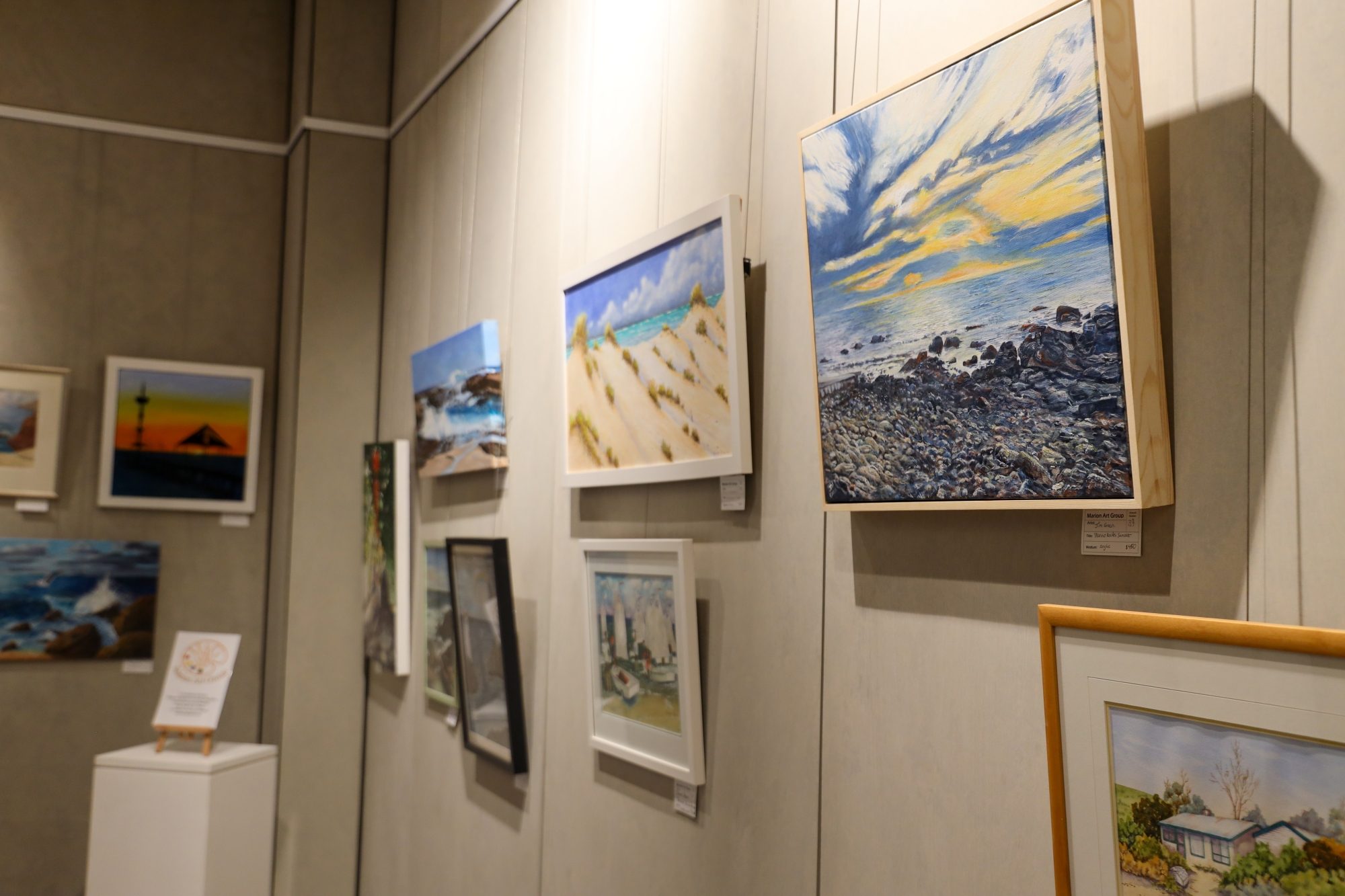 Art gallery wall displaying a variety of framed paintings, including landscapes, seascapes, and beach scenes, under soft lighting.