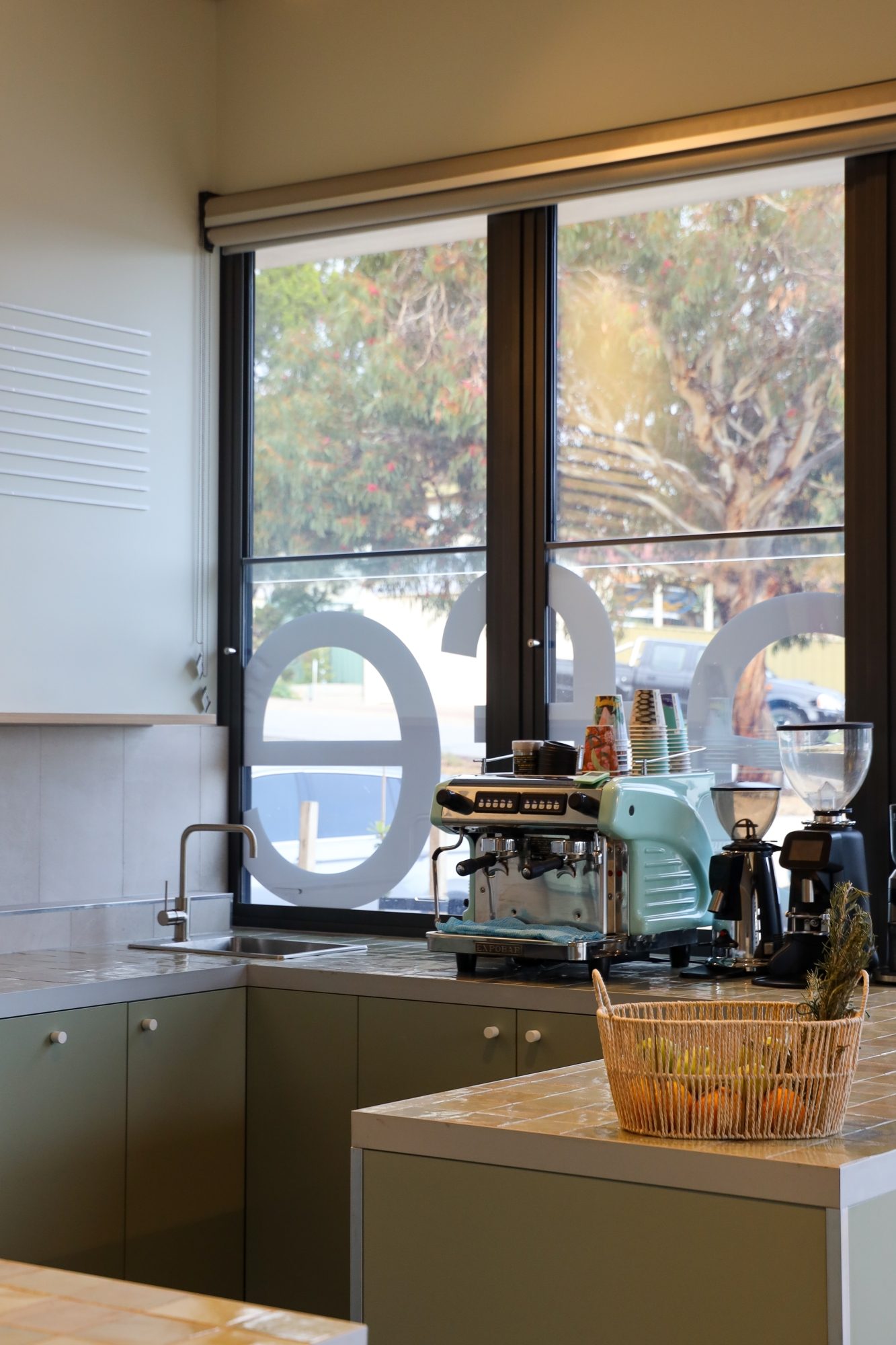 Modern café corner with a coffee machine, grinder, and sink set against a large window with natural light and a view of trees outside.