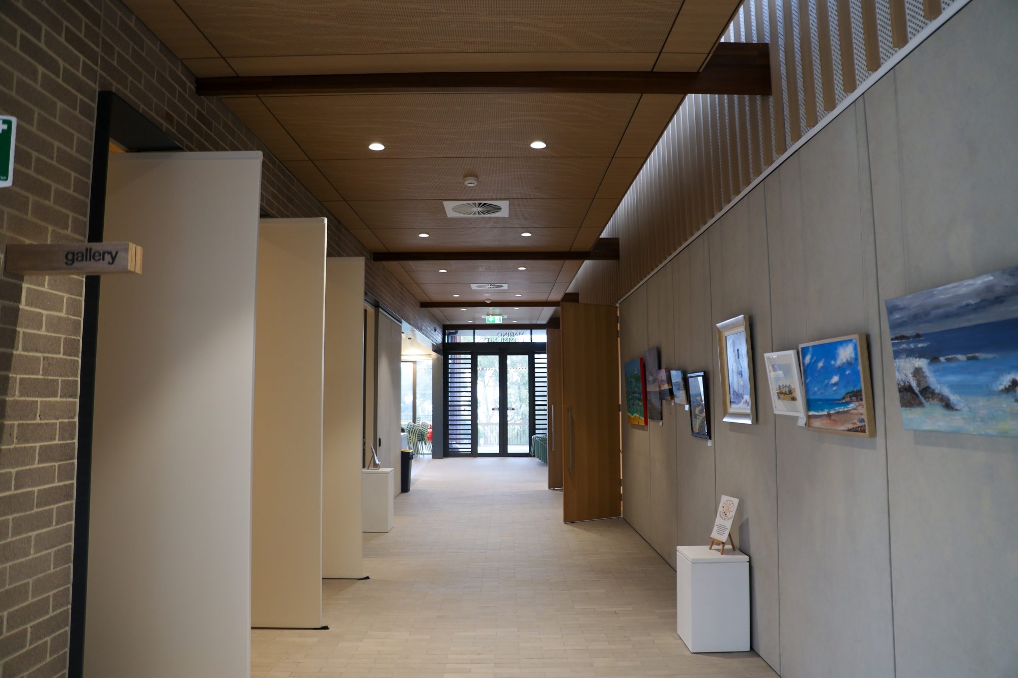 Hallway of a modern building with wooden ceiling, gallery signage, and framed artwork displayed on the wall leading to a glass exit door.