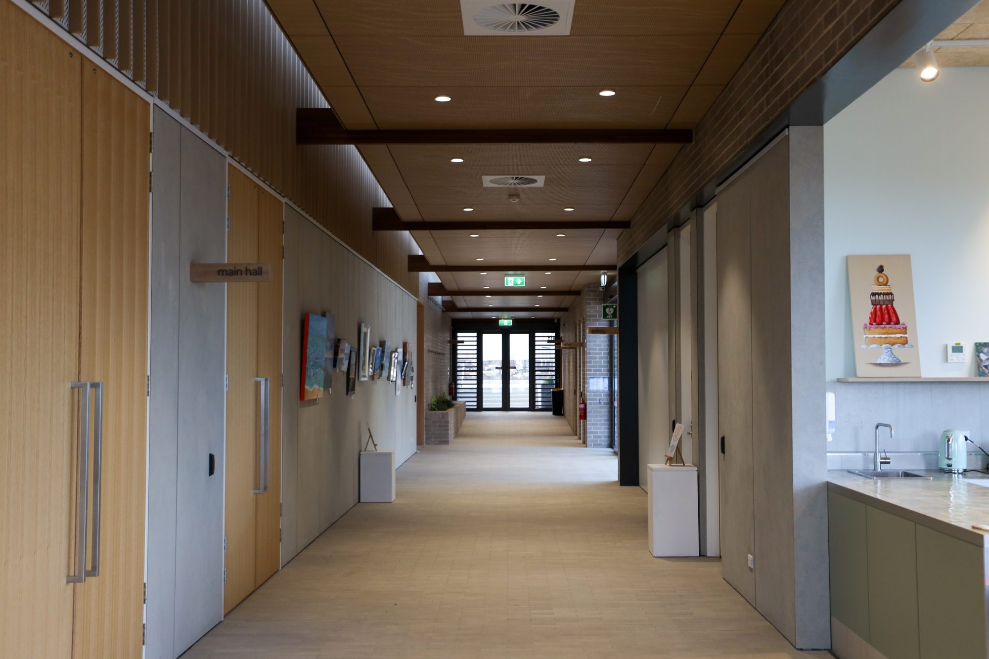 Interior hallway of Marino Community Hall with wood and neutral finishes, decorated with framed artworks on the walls and potted plants near the exit.