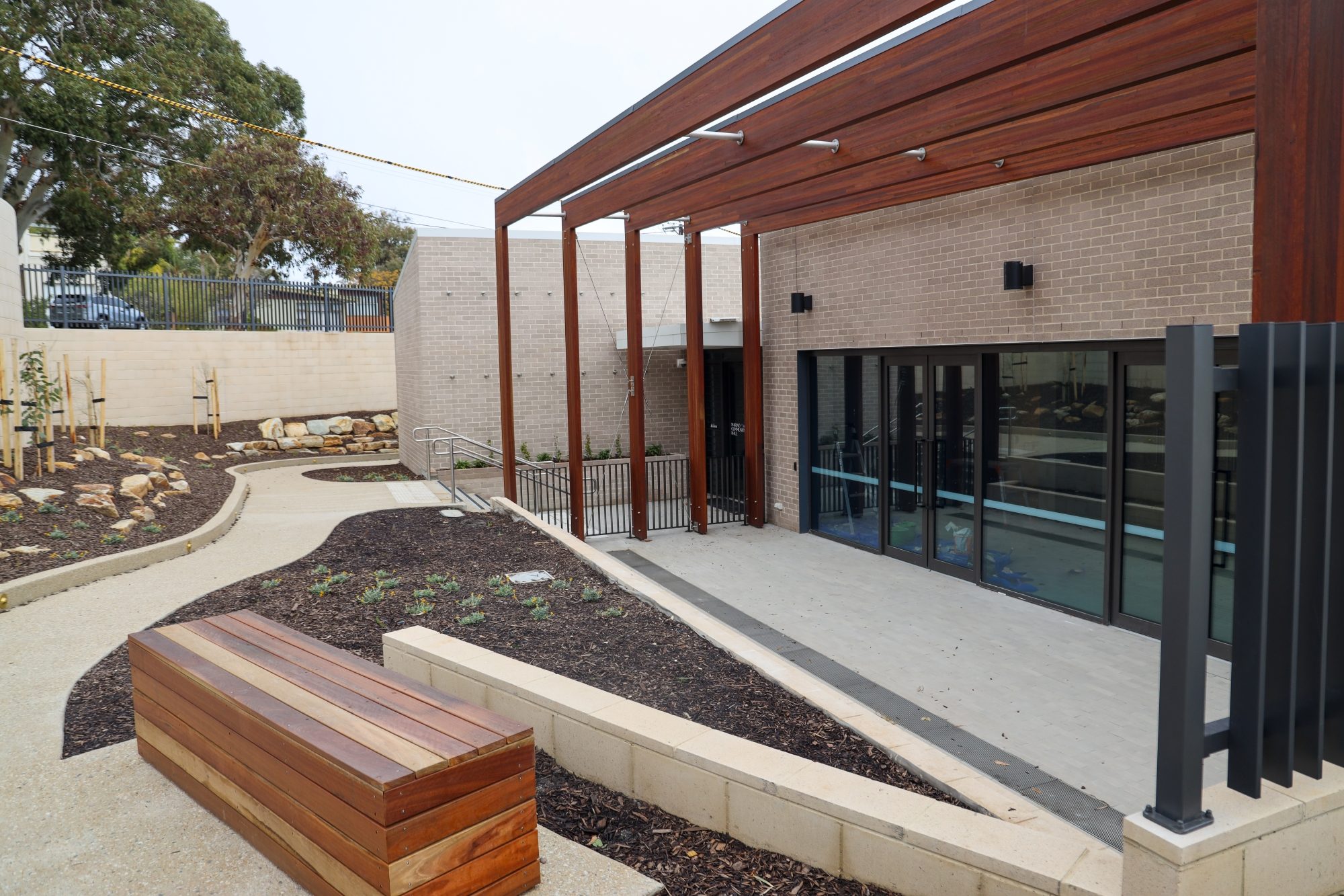 Outdoor area of Marino Community Hall featuring a modern ramp with handrails, wooden seating, landscaped garden beds, and tall timber pergola beams over a paved walkway.