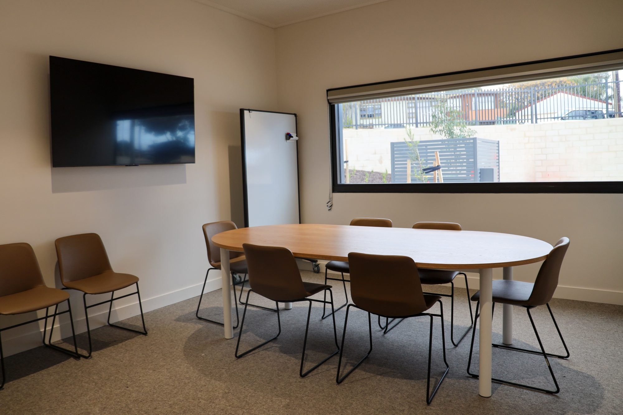 Small meeting room with an oval table, eight chairs, a wall-mounted TV, a whiteboard, and a large window providing natural light.
