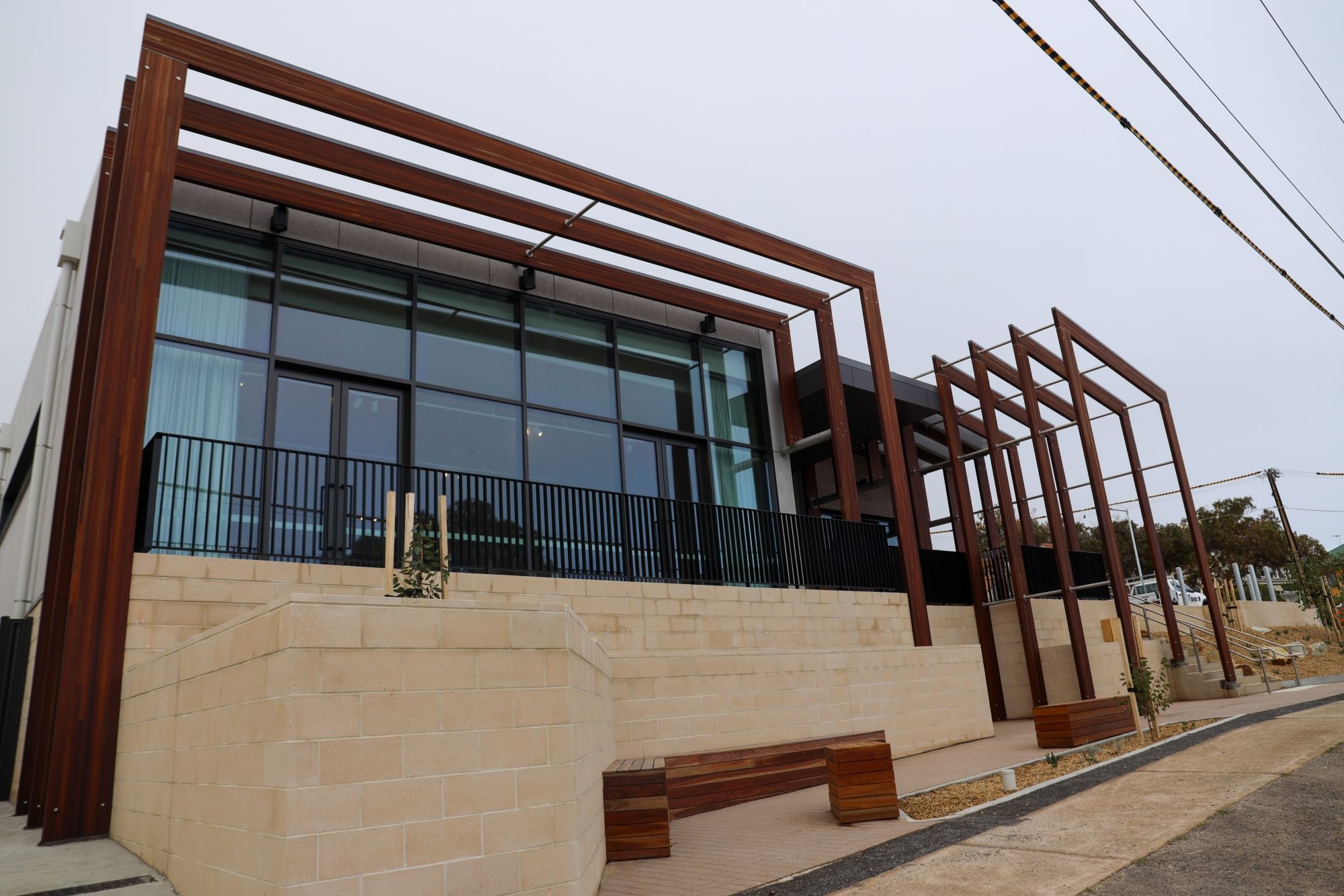 Modern exterior view of Marino Community Hall showing tall wooden architectural frames, large glass windows, stone walls, benches, and a sloped street frontage.