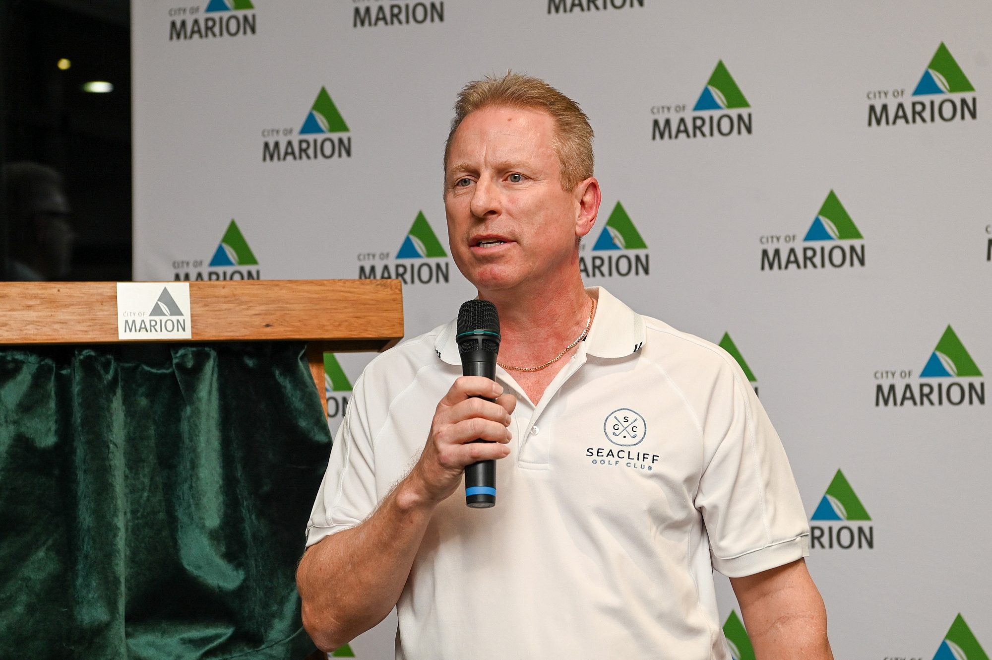 Man in a white polo shirt stands with a microphone in front of a City of Marion branded wall.