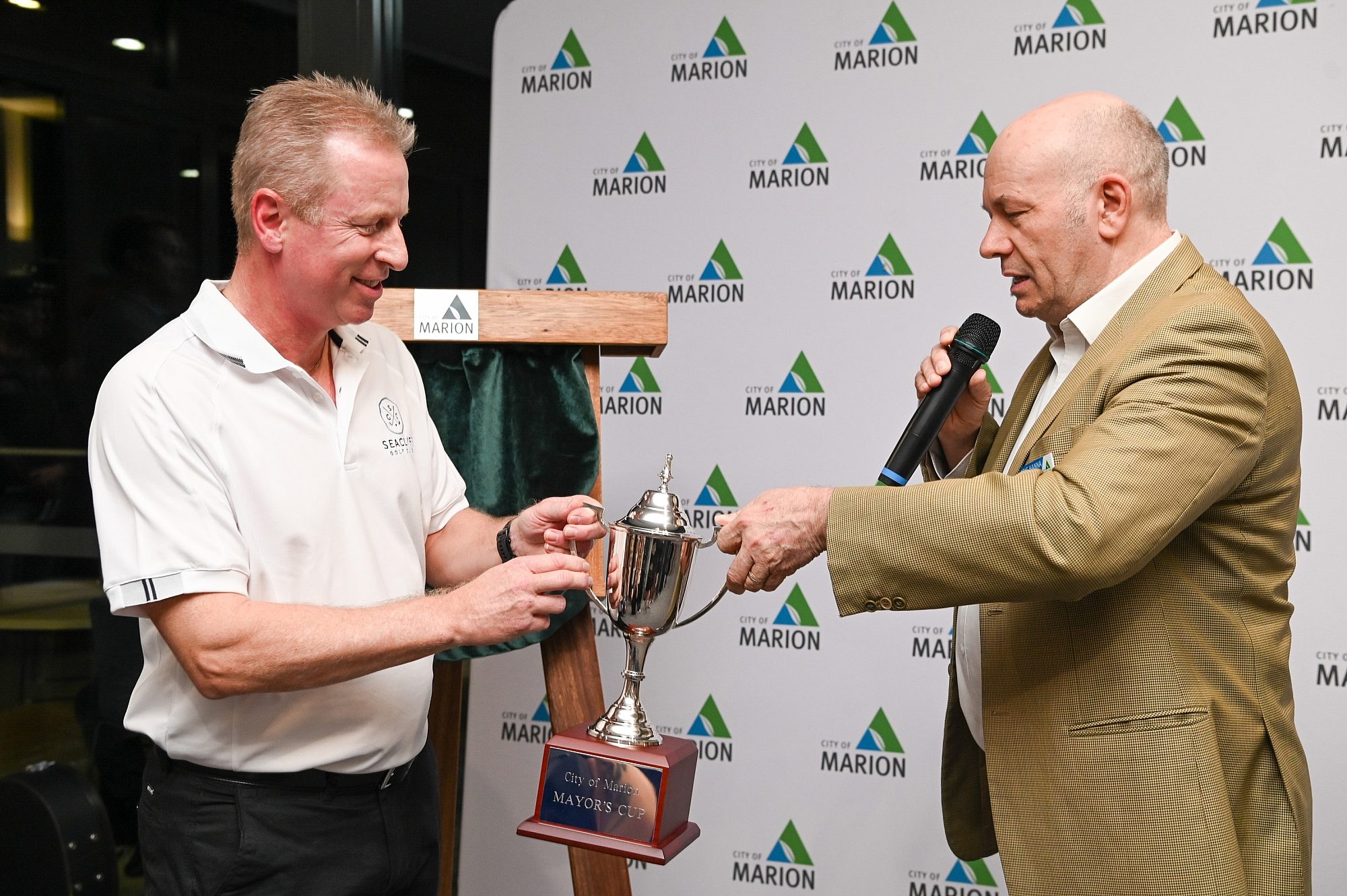 2 men stand in front of City of Marion branded backdrop holding a trophy cup.