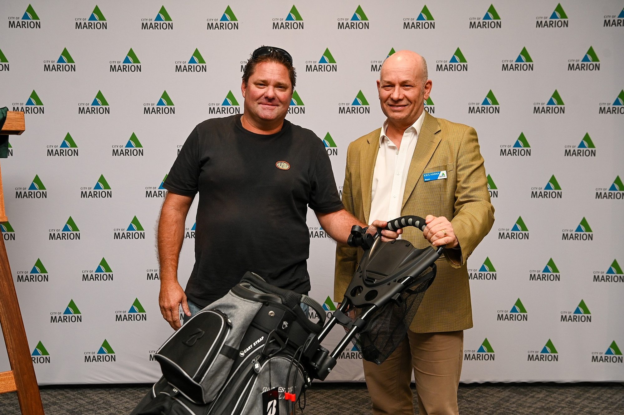 2 men smile at the camera holding a golf bag, in front of a City of Marion branded media wall.