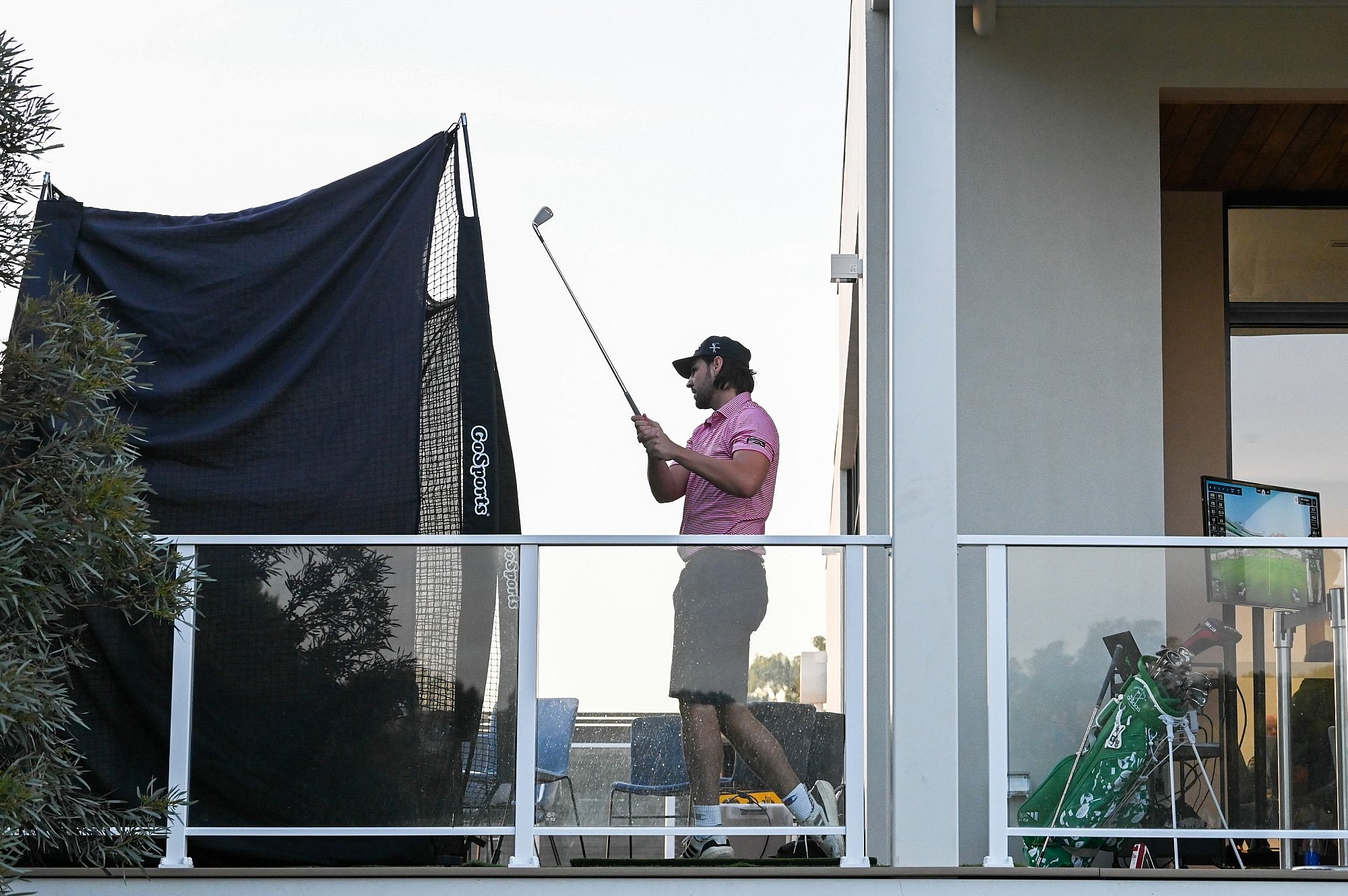 A man practices golf on a balcony, hitting into a GoSports net with a simulator screen and golf bag nearby.