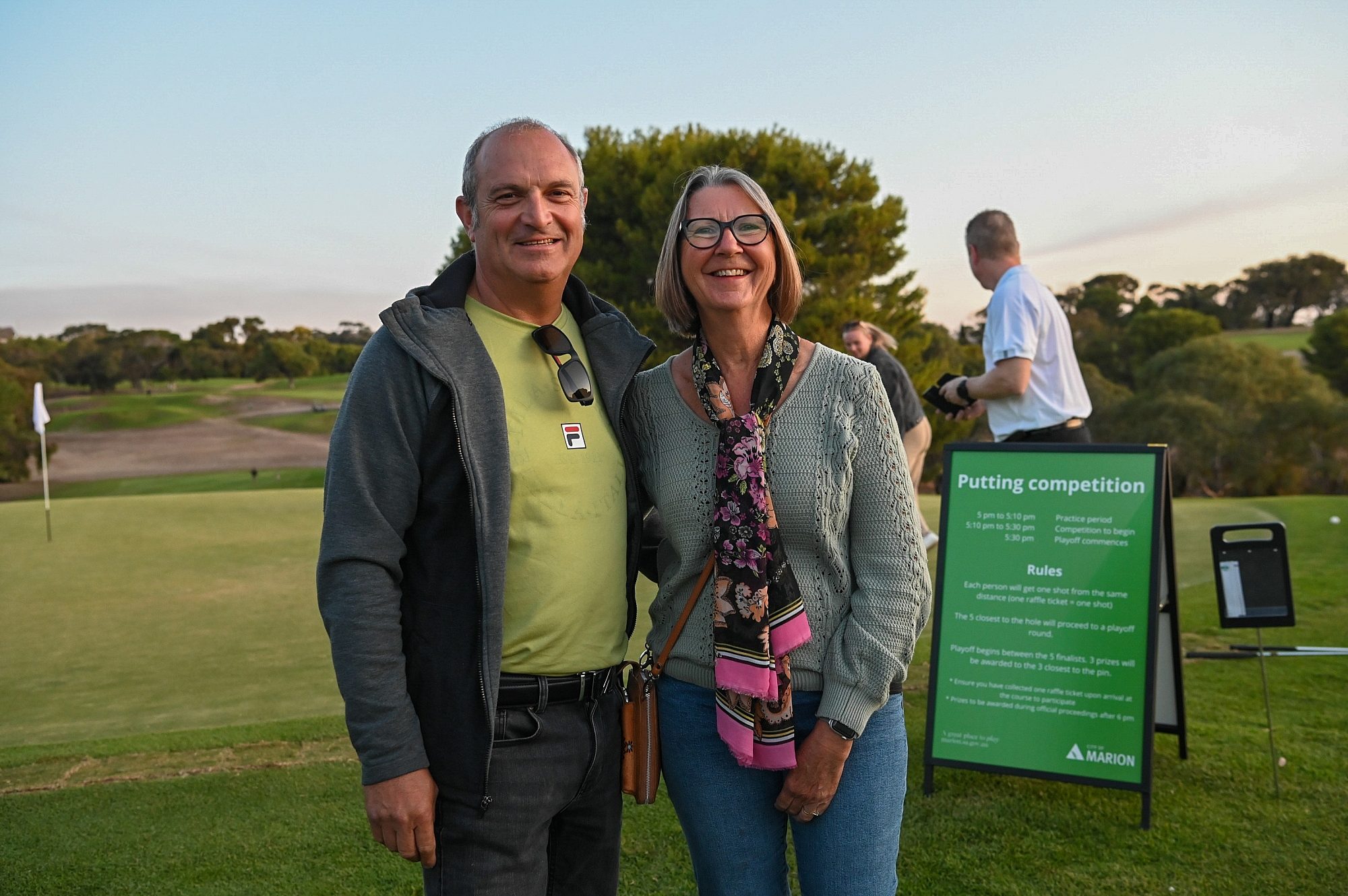 A man and a women smile at the camera, putting competition sign in the background.